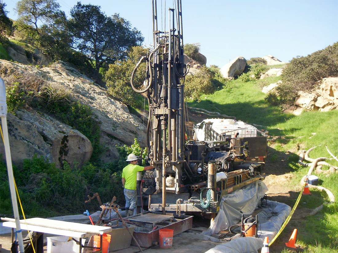 A worker is inspecting a large drilling rig as crews drill a groundwater monitoring well along a roadway surround by green grass, trees and rock formations.