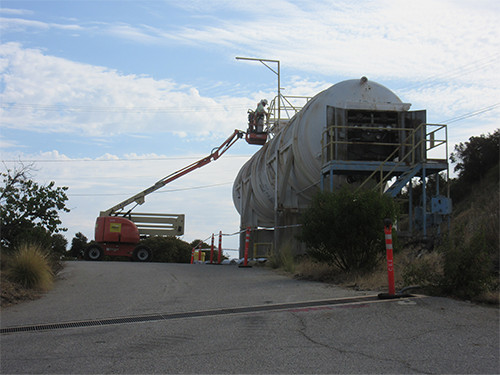 This photo shows a large white tank on the right side of a road. An individual is seen performing abatement activities in preparation for demolition of the tank.