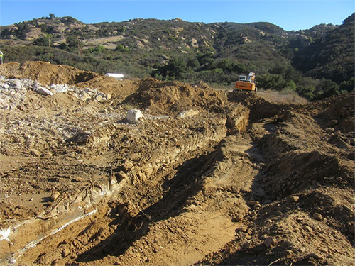 An excavator is shown at the end of a large trench that has been dug several feet into the earth. On the left side of the trench, a large concrete slab is exposed. This is the concrete slab that is being demolished in Phase 4.