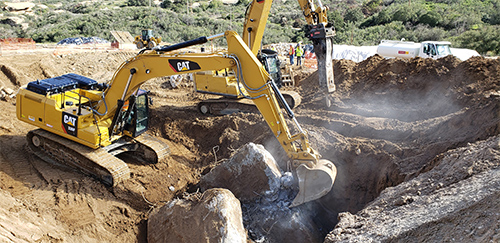 In this photo an excavator is shown digging into a large area several feet into the earth. Much of the concrete slab has been removed but chunks of concrete can be seen on the ground, as well as rebar.