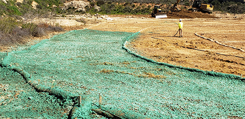 In this photo, the concrete has been removed and the ground has been filled in and smoothed. A worker is shown spraying green hydroseed mix over the surface of the ground to promote revegetation.