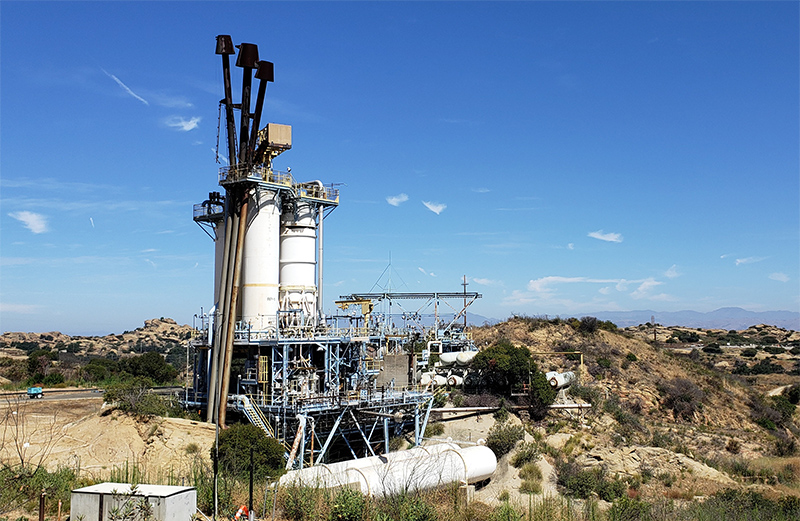 Oct 2020: This photo shows one of the Bravo Test Stands, built into the bedrock. The test stand frame is a light blue and two white tanks stand vertically above the frame. Three black exhaust pipes stick up from the very top of the stand. In the background is sky and mountains in the distance.