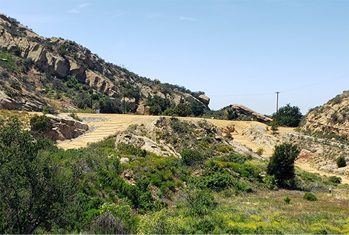 This photo is from a similar angle, after demolition. The same large rock outcrops are in the background. The hillside has been graded and dirt, rock outcrops, and trees and shrubs are all that remain, aside from some straw wattles and strategically placed rocks that are being used for erosion control purposes.