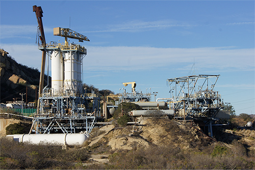 This photo shows the Bravo Test Stands prior to demolition, and fully intact. The stands are blue metal on the bottom, with white tanks stacked on top. Atop the entire stand are large flare stacks that look like exhaust pipes protruding from the top. Large rock outcrops and blue skies are in the background, against which the Bravo Stands are situated.
