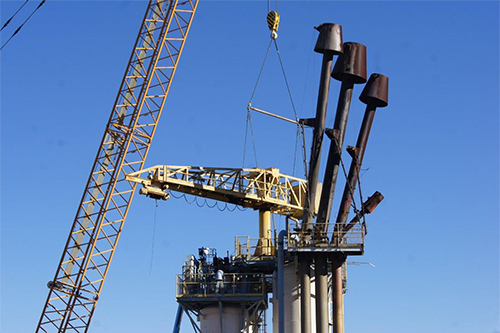 This photo shows a close-up view of the top of Bravo Test Stand 2. The yellow crane on top of the test stand is being pulled away from the stand, and attached to cables of a larger crane that extends up from the ground. The four flare stacks are still intact on top of the test stand and are shown in this photo next to the crane arm being removed from the stand.